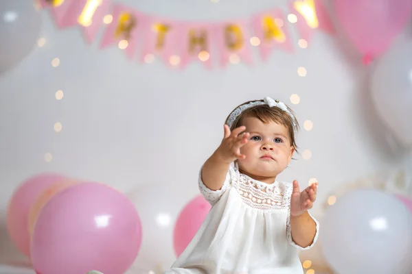First year\'s birthday.a happy little girl in a white tutu dress sits against a background of garlands and pink balloons, celebrating her first birthday. Decoration of the birthday. copy space.