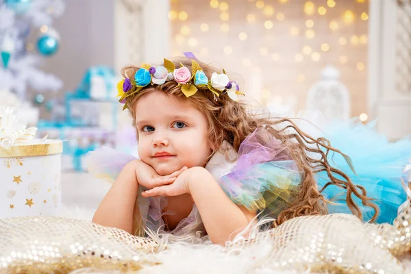 漂亮的小女孩穿着华丽的衣服 头戴皇冠躺在毛绒绒的地毯上 宝宝正在等待假期 并在装饰着白色圣诞树和壁炉的客厅里接受礼物 — 图库照片