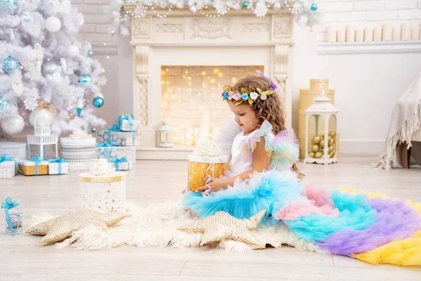 メリークリスマスと幸せな休日 可愛いドレスの女の子ティアラは贈り物を開きます ギフトボックスと暖炉のある木の下の子供 リビングルームにはクリスマスのおもちゃが飾られています 暖かい冬の日 — ストック写真