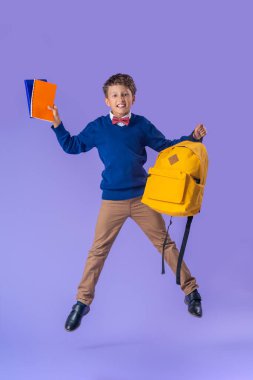 Sırt çantası ve mor arka planda kitapları olan neşeli yaramaz okul çocuğu. Konsept okuluna kadar uzanan dinamik görüntüler. Tatil başlıyor. Okula dönelim. Çocuk çalışmaya hazır..
