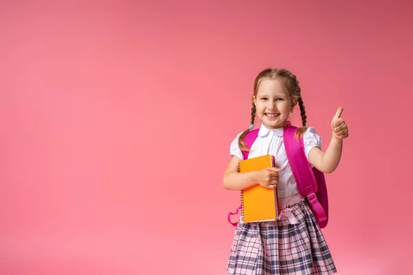 バックパックと本を持つ女の子は 承認の親指のサインを与えます 学校に行くのは初めてです 子供は笑顔でピンクの背景にポーズをとって親指を立てています 学校に戻る — ストック写真