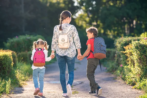 mother accompanies students on road. children with briefcases walk from school, holding their mother in their arms. Parent and elementary school students hold hands. beginning lessons. Autumn.