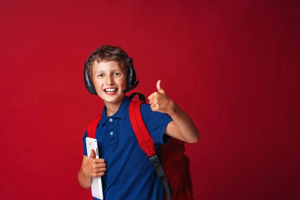 マイクを持ったヘッドフォンの幸せな少年バックパックと赤い背景の彼の手にタブレットコンピュータ 子供は学校に行く準備ができている Eラーニング 距離学習 学校に戻る — ストック写真