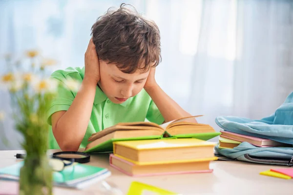 学校の本やノートの山を持つ疲れ果てた少年です 本を持って悲しい少年がテーブルに座って頭を握りしめている 少年は宿題を完成させるのに — ストック写真