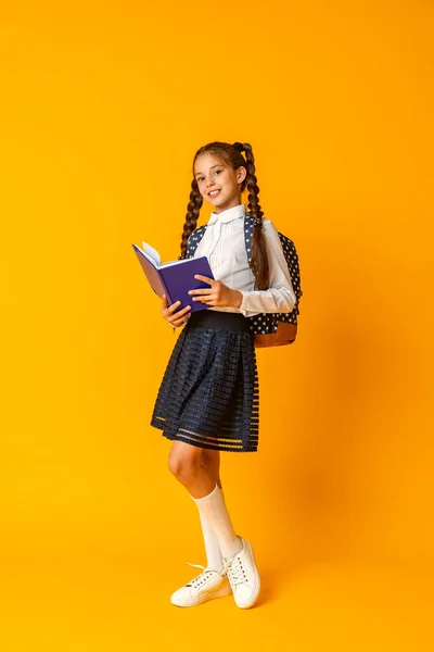 Щаслива Усміхнена Школярка Уніформі Рюкзаком Книга Руках Стоїть Позуючи Жовтому — стокове фото