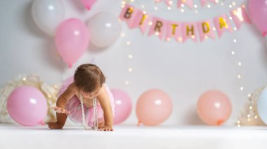 İlk doğum günü. Pembe etekli mutlu küçük bir kız arka planda çelenkler ve pembe balonlarla sürünerek ilk doğum gününü kutluyor. Doğum günü dekorasyonu.