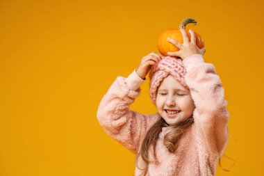 Samimi örgü giysiler ve başlık takan küçük tatlı bir kız elinde küçük bir portakal kabağı tutuyor ve kafasına takıyor. Çocuk gülümser, gözlerini kapatır ve sarı bir arka planda poz verir.