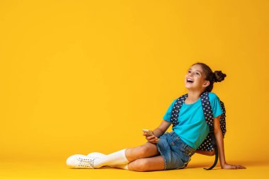 10 yaşında, sırt çantalı, sarı arka planda oturan, elinde cep telefonu olan sevimli, pozitif bir kız öğrenci. Cihazlara ve internete bağımlılık. E-eğitim. Boşluğu kopyala.