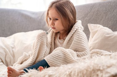 Hasta kız kanepede oturuyor, battaniyeye sarılıp ateşini ölçüyor. Sağlıksız üzgün bir çocuk viral bir hastalık için tedavi edilir, öksürük şurubu. Salgın koronavirüs, covid-19..