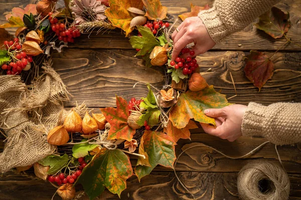 Kırsal ahşap tahtalarda sonbahar çelengi renkli yapraklar ve doğal malzemeler yapmak. Üst görüşlü kadınların elleri, kahverengi ahşap masa üzerinde sonbahar hasat ve yapraklarını yuvarlak çelenk yapar. İç mekan için dekorasyon.