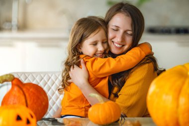 Sevimli küçük kız annesine sevgiyle sarılıyor. Kız ve anne masaya oturur ve şefkatle kucaklaşırlar, gözlerini kaparlar, parlak güzel balkabaklarının yanında. Annenin sevgisi ve bakımı.