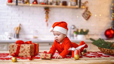 Kırmızı kostümlü bebek, tahta yüzeyde Noel süsleriyle yatıyor. Bebek, Noel kutlaması sırasında hediye kutularının yanında karnının üstünde yatıyor. Etrafındaki dünyayı dikkatle incele. Noel arifesi