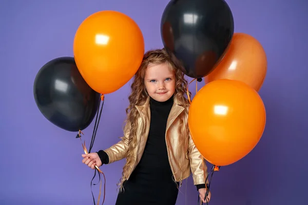 时尚的小女孩 穿着时髦的金色夹克 手里拿着橙色和黑色气球 站在那里 看着相机 微笑着 隔离在紫色背景上 — 图库照片
