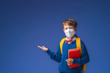 Koruyucu maskeli okul çocuğu, üniformalı, mor arka planda kitap ve sırt çantalı. Heyecanlı çocuk elini yana doğru uzatır ve dikkatini boş alana verir. salgın koronavirüs