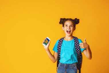 10 yaşında, sırt çantalı, sarı arka planda oturan, elinde cep telefonu olan sevimli, pozitif bir kız öğrenci. Cihazlara ve internete bağımlılık. E-eğitim. Boşluğu kopyala.
