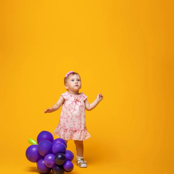 可爱的小宝宝躺在黄色的背景上 带着葡萄形状的气球 令人惊讶的小女孩在工作室的一面黄色的墙的后面看着旁边 复制空间 — 图库照片