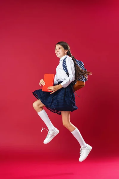 淘气的女学生 穿着校服 背着背包 背着书跳到红色的背景上 动态图像 回到概念学校去 假期开始了回学校去3 女孩子准备好要学习了 — 图库照片