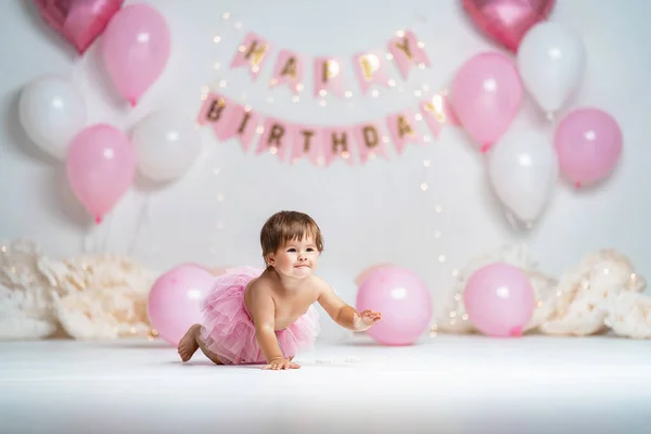 First year\'s birthday.a happy little girl in a pink tutu skirt crawls on a background with garlands and pink balloons, celebrating her first birthday. Birthday decoration.