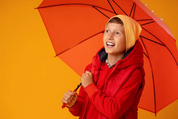 穿着秋天衣服的快乐喜庆的男孩 在黄色的背景下 在橙色的伞下笑着 孩子高兴地微笑着 把目光移开了 秋天的概念 温暖的秋天衣服 — 图库照片