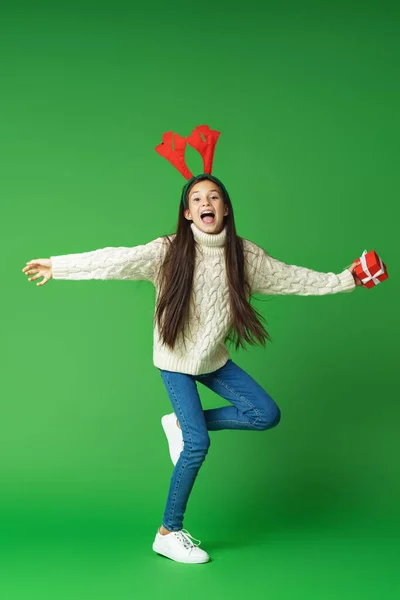 一个快乐快乐的女孩穿着针织毛衣 手里拿着一个盒子的大鹿角作为圣诞礼物的画像 一个孩子在绿色的背景上跑着跳着 动态图像 复制空间 — 图库照片