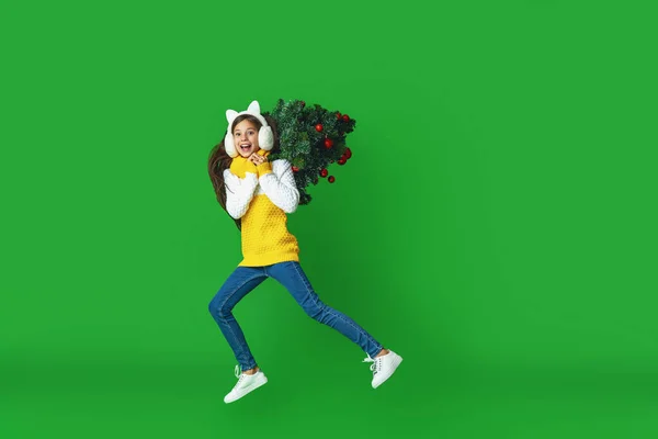 身穿针织毛衣 头戴毛皮耳机 头戴圣诞树 肩上挂着红色圣诞彩球的快乐少女 走在绿色的背景上 动态图像 — 图库照片