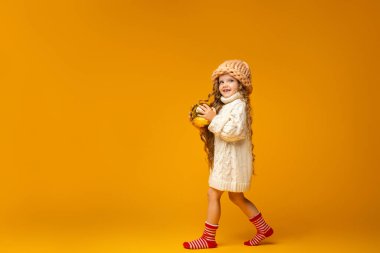 6 yaşında, Noel çoraplarıyla örgü örmüş küçük beyaz bir kız elinde büyük kırmızı bir Noel topuyla Stüdyo 'da sarı arka planda yürüyor. Yeni yıla hazırlanıyorum. Boşluğu kopyala.
