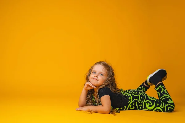 可爱的高加索女孩6岁 卷曲的头发穿着时髦的衣服躺在工作室的黄色背景上 那孩子笑了笑 用手捂住头 复制空间 — 图库照片