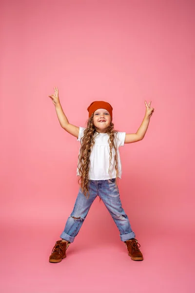 时尚的小女孩 卷曲的头发 戴着时髦的帽子 撕破的牛仔裤 全身上下都站着粉色的背景 显示胜利的标志 小孩无牙地微笑着 情感形象 — 图库照片