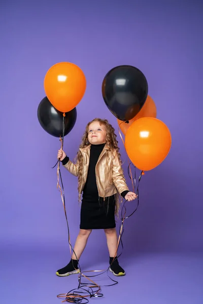 时尚的小女孩 戴着太阳镜 卷曲的头发 穿着时髦的衣服 手里拿着橙色和黑色的气球 高高地站在那里 望着紫色背景的气球 — 图库照片