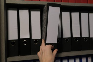 Dosyalar, düzen, dosyalama ve işyeri organizasyonu kavramları ile dolu bir raftan boş etiketli siyah bir ofis klasörünü çeken bir elin yakın çekimi