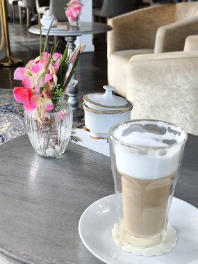 Bir kafedeki bir masanın üzerinde yumuşak süt köpüklü bir bardak sıcak kahve, yanında da narin pembe çiçekli bir vazo ve şeker kasesi var.