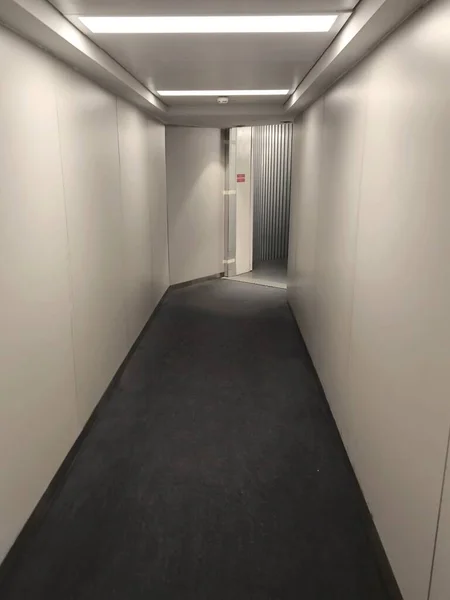 Yolcular modern bir havaalanı terminalinde uzun ve parlak bir koridorda yürürler. Büyük pencerelerden akan güneş ışığı yerde ve duvarlarda uzun gölgeler oluşturarak mimari çizgileri aydınlatıyor..