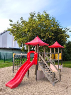 Bright and modern playground surrounded by green trees. Play complex with a red slide, ladder and rope net