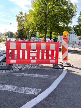 The photo shows road repair work during daytime on a city street. Bright red and white plastic barriers with yellow signal lights installed on the paved road