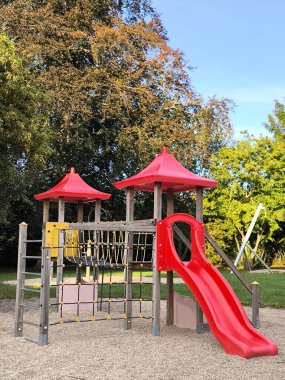 Bright and modern playground surrounded by green trees. Play complex with a red slide, ladder and rope net