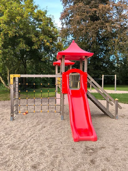 Bright and modern playground surrounded by green trees. Play complex with a red slide, ladder and rope net