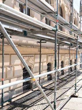 The facade of a historic building, partially hidden behind scaffolding. Metal pipes and platforms create a complex geometric pattern.