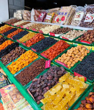 Bir markette kutulara konmuş renkli kurutulmuş meyve çeşitlerinin yakın plan çekimi. Çerçevede hurmalar, kurutulmuş kayısılar, koyu ve açık üzüm, kuru erik ve diğer kuru meyveler görülebilir. Doğal tatlıların bolluğu ve çeşitliliği