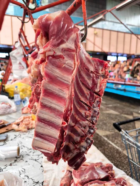 Fresh pork ribs hanging on a hook at a meat market counter. The crisp lines of the bones and layers of meat indicate the high quality of the product. The photo conveys the realistic atmosphere of a local market.