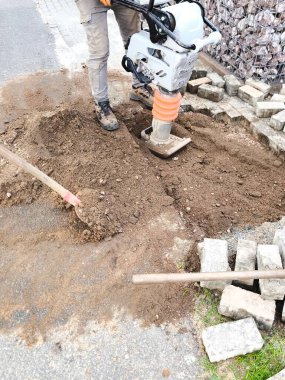 A man in work clothes and gloves uses a vibratory rammer vibratory rammer to compact the ground above a construction site.