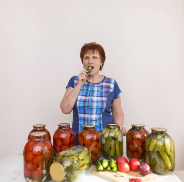 bir mavi Kareli tunik kadında tablosunun yanındaki mutfakta bir salatalık var. masanın üzerine kavanoz turşu, salamura domates ve salatalık, masanın üzerine taze domates, salatalık, soğan, sarımsak, sebze, yemek