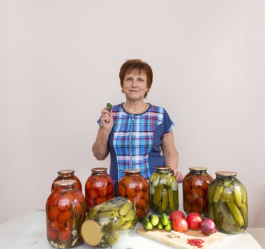 bir mavi Kareli tunik kadında tablosunun yanındaki mutfakta bir salatalık var. masanın üzerine kavanoz turşu, salamura domates ve salatalık, masanın üzerine taze domates, salatalık, soğan, sarımsak, sebze, yemek