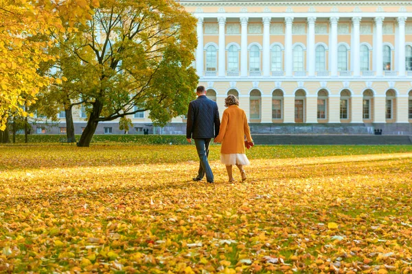 couple walks through the autumn foliage across the glade against the background of yellow fallen leaves against the background of the building, wedding in autumn, bride and groom