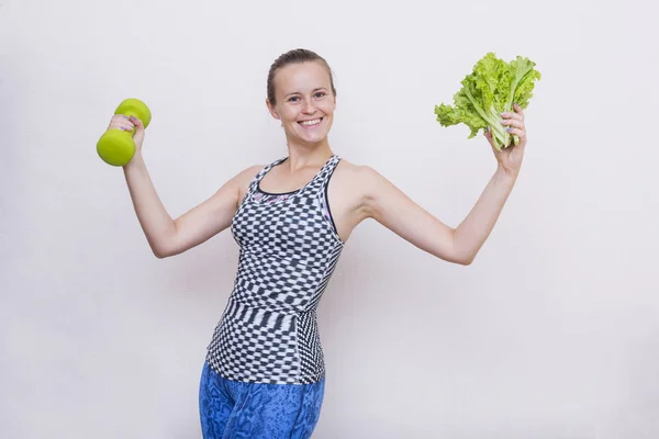 white girl in a sport  top and leggings laughs,  girl holds a dumbbell and a bunch of green salad in her hands, girl with dumbbells,  young woman smiles