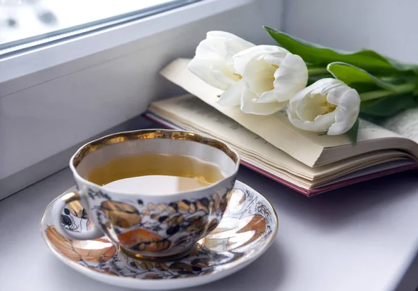 Cup of tea on the windowsill, open book, 3 white tulips, flowers