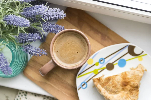 Cup of coffee with milk, a piece of cake on a plate. blue lavender branch on the windowsill