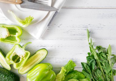 Yeşil biber, salatalık, yeşil marul yaprakları, ıspanak, roka, bıçak, çatal, beyaz bir havlunun üzerindeki beyaz havlu, ahşap arka plan, üst manzara, yeşil sebzeler ve otlar.