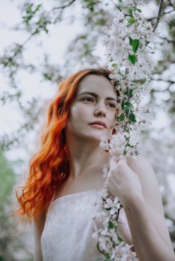 Bahçedeki bir elma ağacının pembe çiçekleri arasında pembe elbiseli 1 beyaz genç kadın.