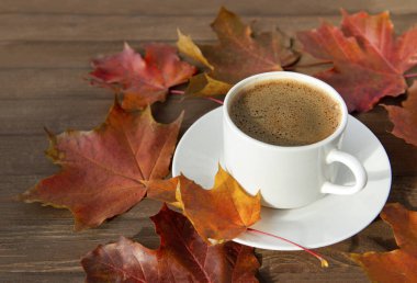 Bir fincan sade kahve, çay tabağı, kestane rengi sonbahar akçaağaç yaprakları kahverengi ahşap arka planda.
