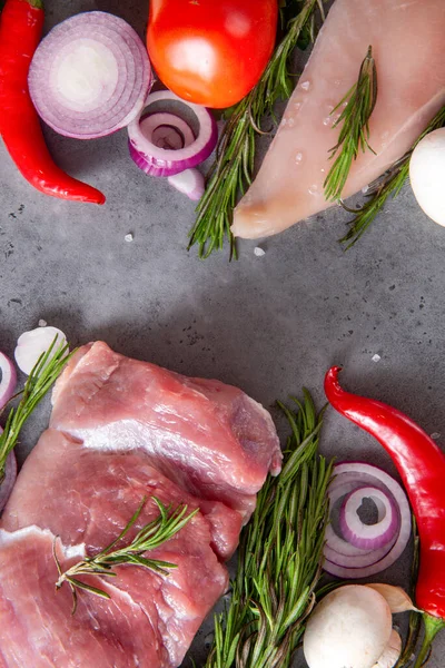 pieces of raw chicken fillet, pork tenderloin, meat, vegetables, herbs, tomato, onion, rosemary, chili pepper, mushrooms on a gray background